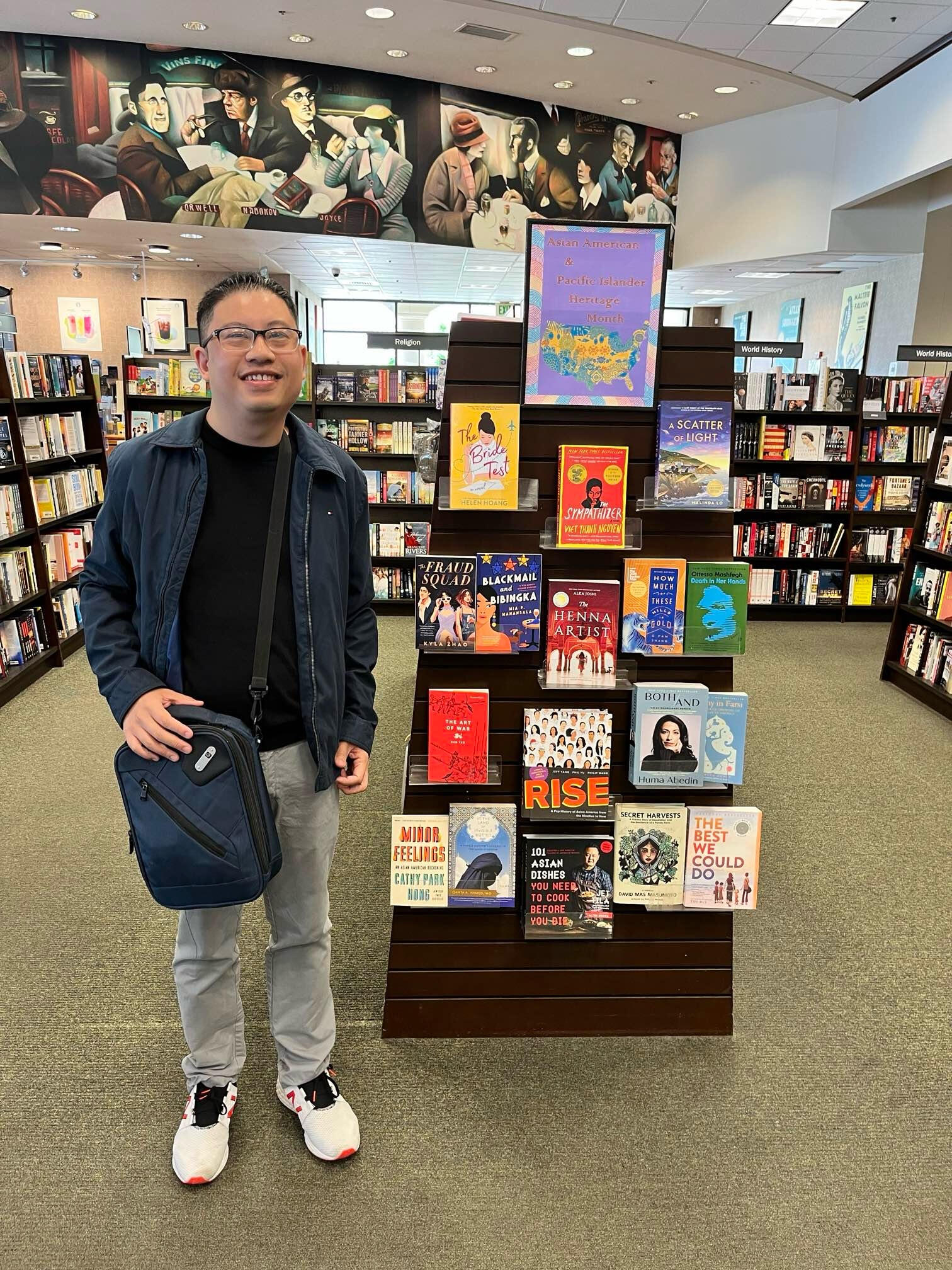 Inside a book store, Martin, a Vietnamese American man with black hair, wearing glasses, a blue jacket, black shirt, gray pants, and white shoes, smiles in front of a book display with an array of Asian American authors.