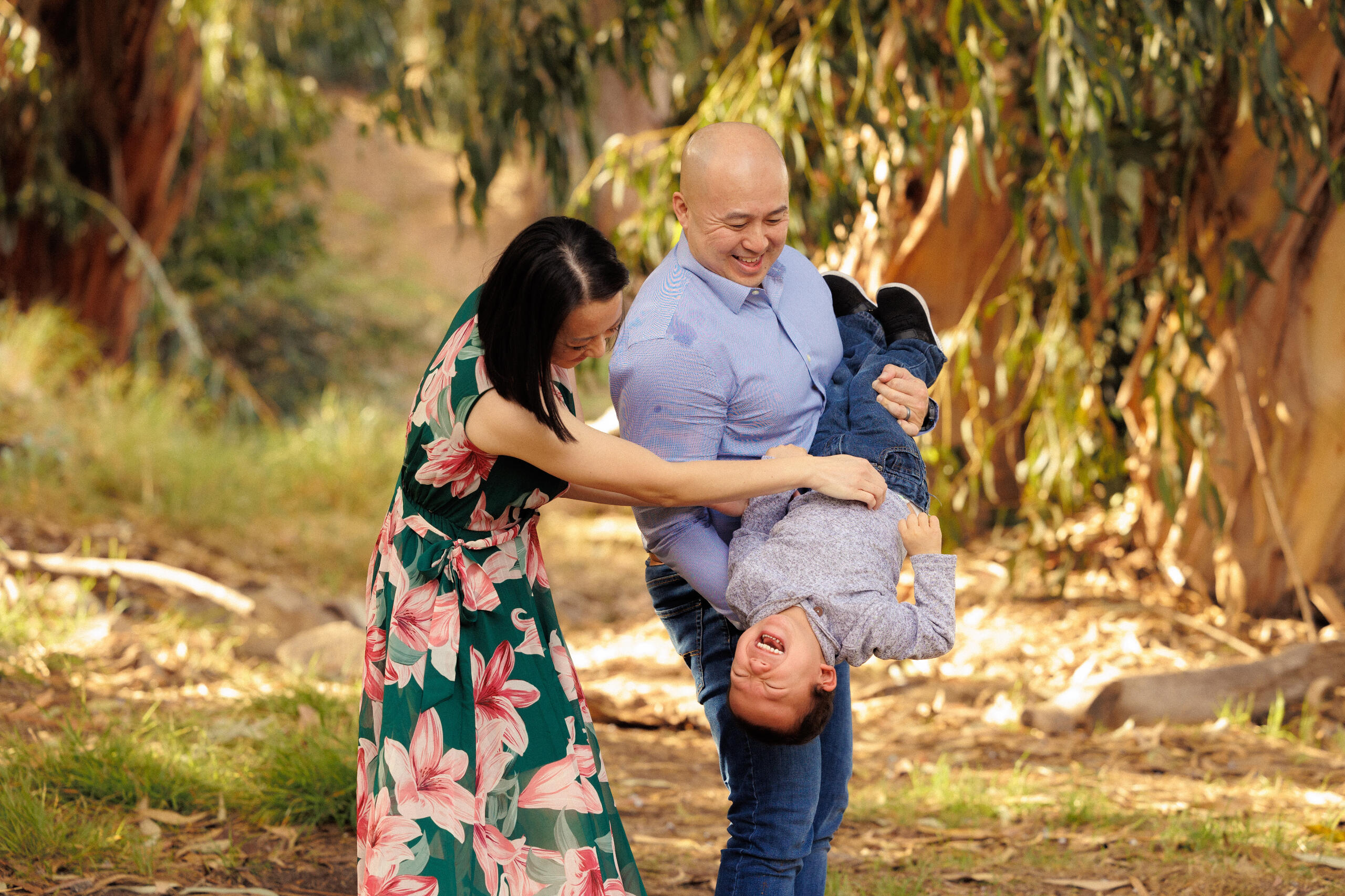 A photo of Irene Nham, her husband, and son smiling together in the moment as they're having fun with a backdrop of forest park at sunset.